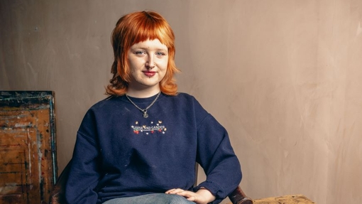 A pic of a student in a blue top and jeans sitting on a chair next to a table.