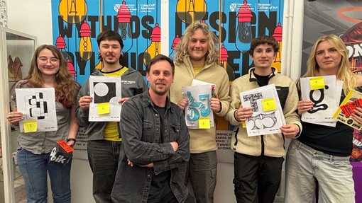 5 students posing in front of a wall that reads 'Design Week 2026', each holding up pieces of their work. A man stands in the middle of the students crossing his arms and looking proud.