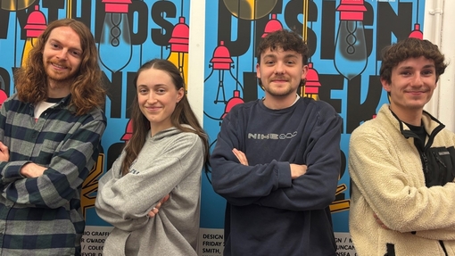 4 students stood in front of wall with graphic that says 'Design Week 2026'. Each one is crossing their arms, looking proud.