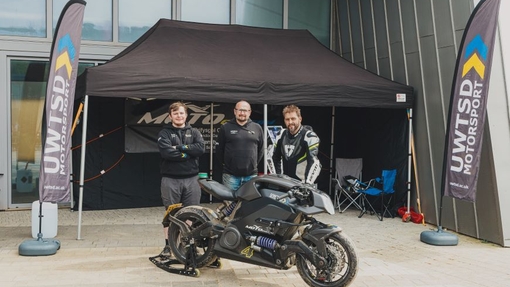 Three men standing in front of a motorbike with a branded gazebo in the background.