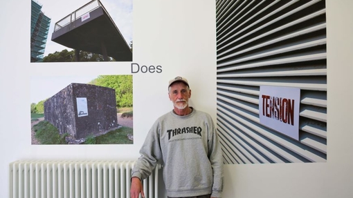 A man standing in front of a white wall, with pictures of his art work.