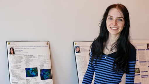 A student dressed in a blue top standing proudly and smiling in front of a board exhibiting her project.