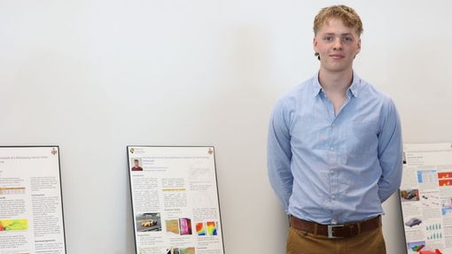 A student dressed in a blue shirt standing proudly by an exhibition of his work.