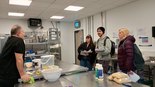 Three students in a catering kitchen at the university talking to a chef