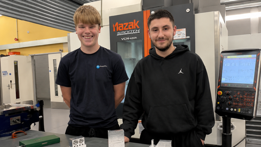 Two apprentices in dark coloured clothing standing in a workshop.