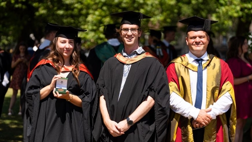 A garduate and two lecturers smiling and standing proudly in their caps and gowns.