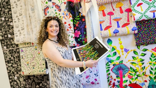 A smiling graduate surrounded by colourful creative work.