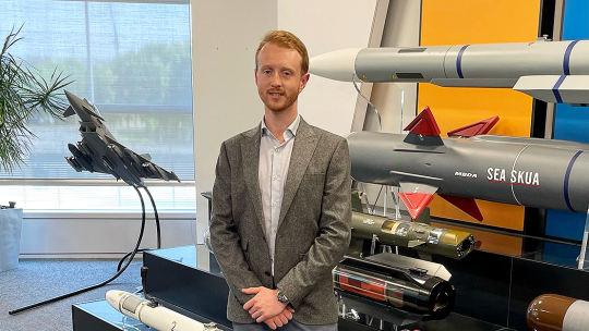 Wearing a grey jacket over an open-necked shirt, Max Hart stands in front of a display of missile casings, including a Sea Skua.
