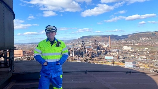 Wearing a hard hat and fluorescent overalls, Daniel Richards stands on the roof of a cooling tower; the Port Talbot steelworks and hills behind Briton Ferry form the background.