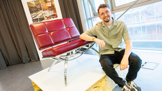 A smiling student, seated next to the chair, with his designs on a board in the background.