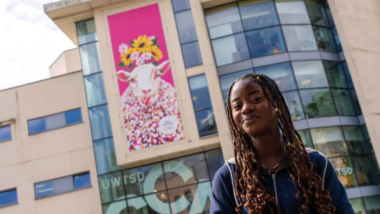 A smiling student standing outside a building which displays her colourful banner image.