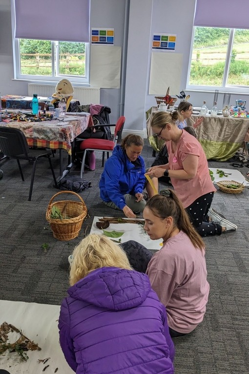 Cyfranogwyr yn eistedd ar lawr dosbarth yn gweithio gyda dail a brigau a gasglwyd yn yr awyr agored.