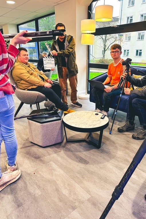 A group of students filming: one holds a clapperboard; another a boom microphone; another adjusts the camera.