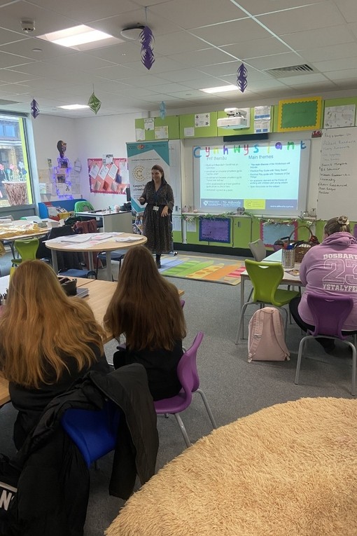 Mae darlithydd mewn ystafell ddosbarth wedi’i haddurno’n lliwgar yn siarad o flaen sleid dwyieithog sy’n cael ei daflunio ar fwrdd gwyn.