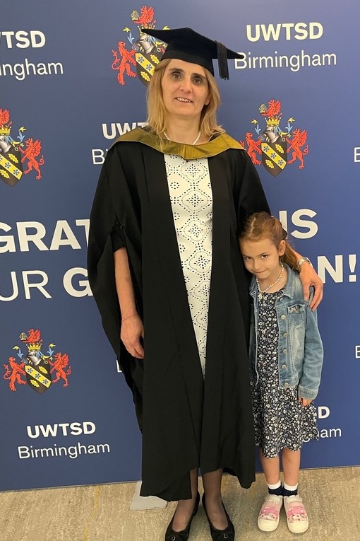 Gan wisgo gwisg academaidd, mae Oslava Chougleva yn gwenu i’r camera; mae un llaw yn gorffwyso ar ysgwydd merch ifanc, sef ei merch.