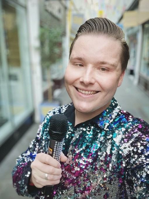 Wearing a besequined shirt and holding a microphone, Jake Sawyers smiles to the camera..