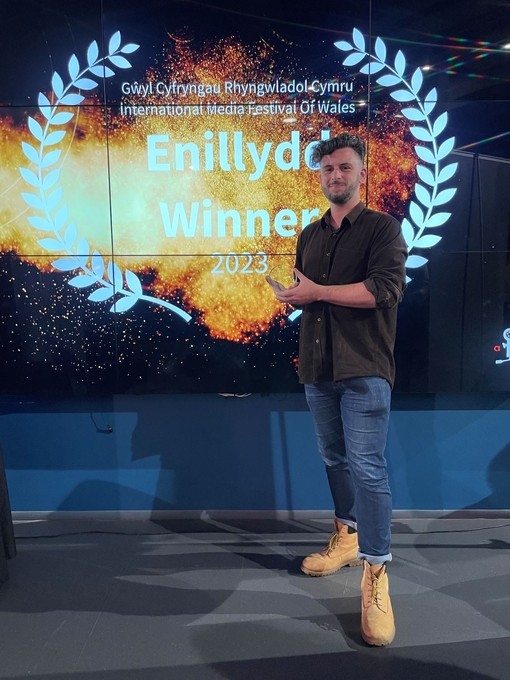 Rhys Cannon yn dal ei wobr; y tu ôl iddo ar sgrin mae coron wen o ddail llawryf yn amgylchynu’r geiriau Enillydd Winner 2023.