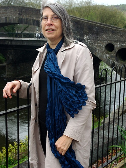Yn gwisgo sgarff shiffon las a chot law lwydfelyn, mae’r Athro Jane Aaron yn pwyso yn erbyn rheiliau wrth lan afon; y tu ôl iddi mae pont garreg fawr yn arddull y bedwaredd ganrif ar bymtheg.