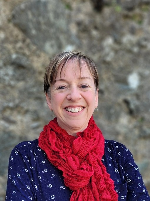Mae Dr Saran Elin Roberts yn gwenu’n llydan ac yn gwisgo sgarff goch dros dop glas.