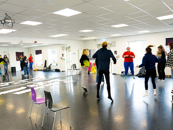 A circle of seven students jump in synch in a dance rehearsal studio with mirrored wall.