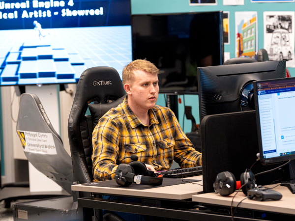 A student sits upright at a PC; behind him, a large screen reads: Unreal Engine 4 Technical Artist – Showreel. 
