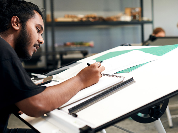 A student concentrates on his sketchbook, which is supported by a large drawing board.