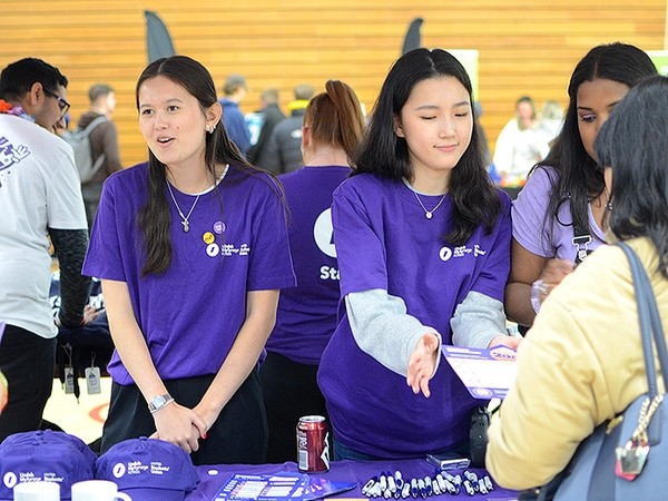 Two young women in purple Student Union talk to people browsing a stall displaying Student Union merchandise.