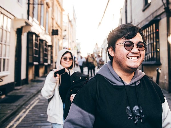 A young man wearing round shaded glasses grins as he walks down a cobbled street.