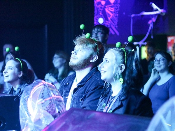 In a busy room lit with purple light, a young man and woman wearing green antennae head-boppers look up towards something off-camera.