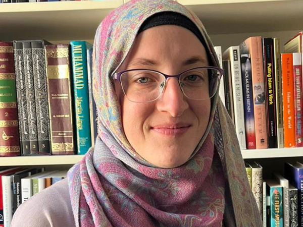 Wearing a pale pink hijab and glasses, Dr Laura Jones looks towards the camera and smiles; the books on the shelves behind her include titles on Islamic theology.