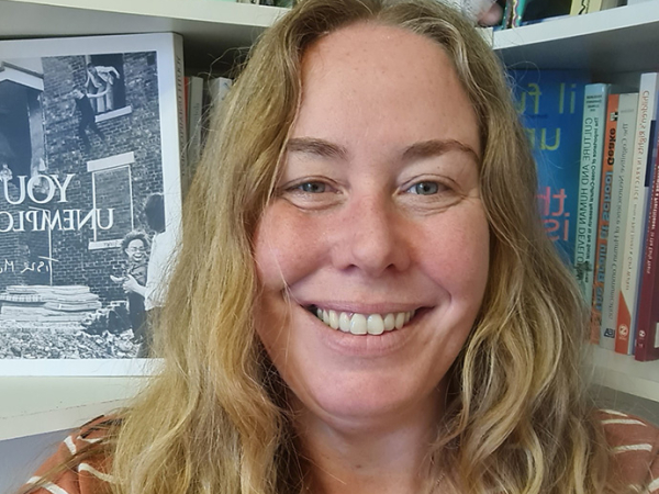 Standing in front of packed bookshelves, with long blonde hair and wearing a silver heart-shaped locket, Dr Jessica Pitman smiles into the camera.