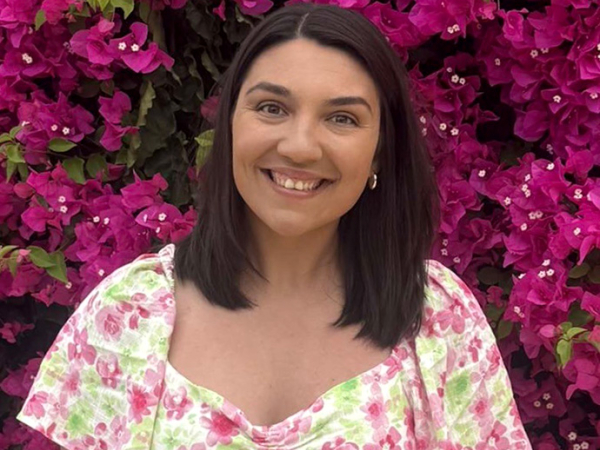 With long dark hair and wearing a floral summer dress, Natasha Jones stands smiling in front of a flowering bougainvillea bush. 