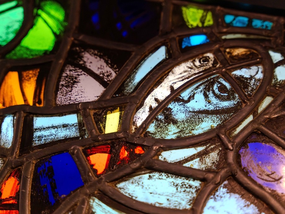 Close-up on a stained glass panel showing a serious human face divided in two by a strip of lead; the face is almost monochrome, but surrounded by pieces of glass in gemstone greens, blues and oranges.