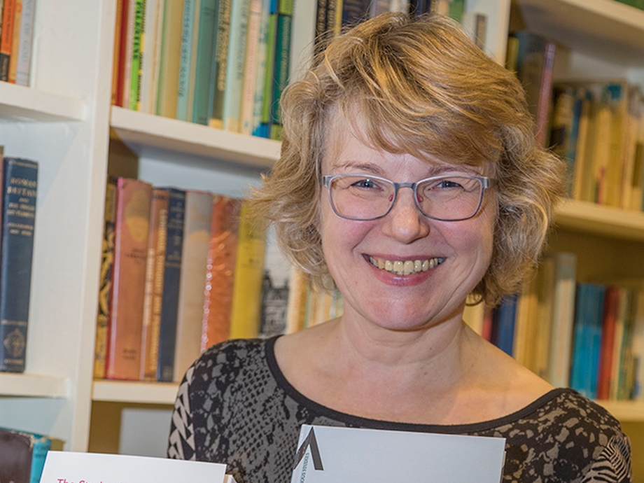 Standing in front of a bookcase, Professor Bettina Schmidt smiles towards the camera while holding up two of her own works: The Study of Religious Experience – Approaches and Methodologies; also Spirits and Trance in Brazil. 