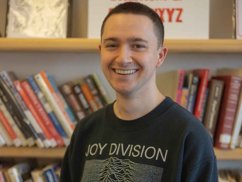 A smiling student wearing a black sweatshirt standing in front of a bookshelf full of books.