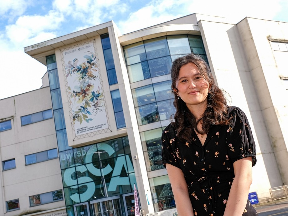 A smiling student standing in front of a giant canvas on the top of a University building in Swansea which features her work.
