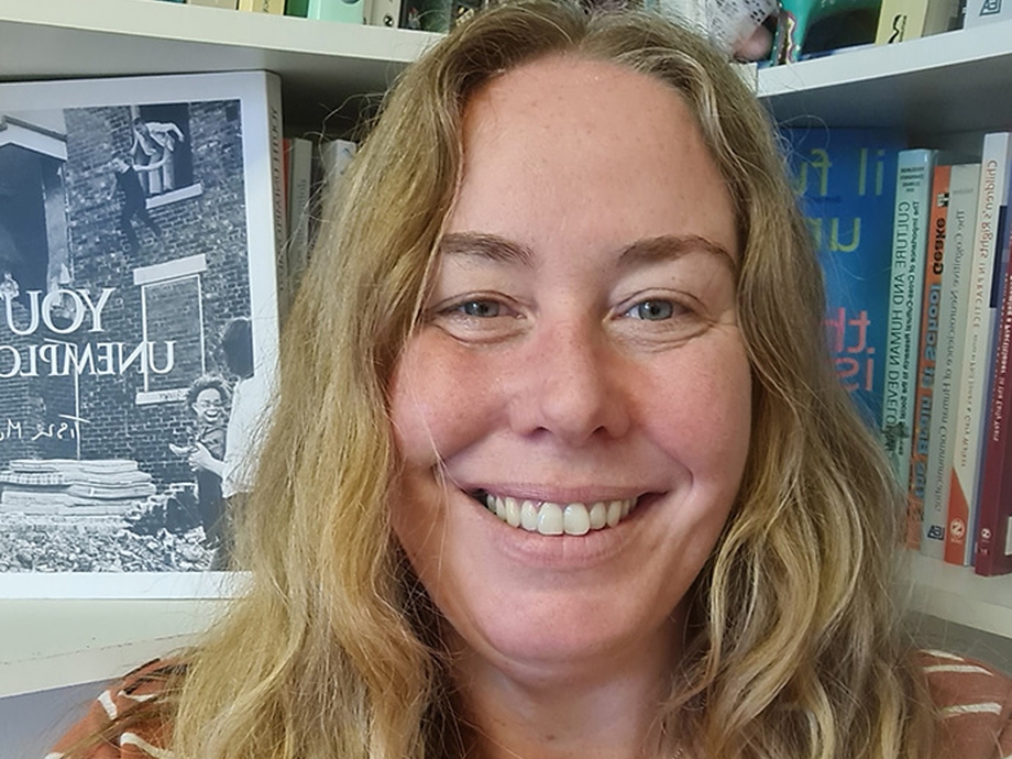 Standing in front of packed bookshelves, with long blonde hair and wearing a silver heart-shaped locket, Dr Jessica Pitman smiles into the camera. 