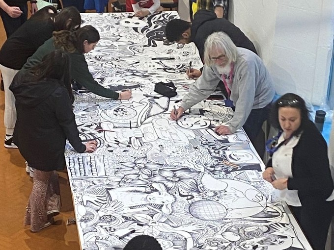 Students, refugees, and asylum seekers drawing on a large piece of paper with black pens, to create a large collective artwork.