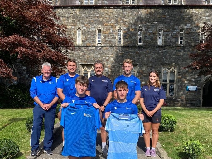 Staff a myfyrwyr yn gwisgo crysau-t chwaraeon glas ac yn dal rhai i fyny i’r camera eu gweld.
