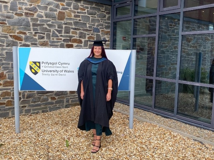 Yn gwisgo gŵn a het academaidd, mae Zoe Vaughan yn sefyll yn gwenu y tu allan i Ysgol Fusnes Caerfyrddin.