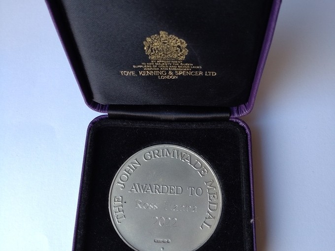 Medal arian-lwyd yn cael ei harddangos mewn blwch â leinin du; mae’r geiriau ar y fedal yn darllen – The John Grimwade Medal Awarded to Ross Hanna 2022; o dan warant frenhinol mae’r testun aur ar leinin y blwch yn darllen –Toye, Kenning and Spencer Ltd London.