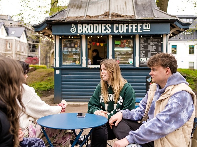Eistedda pedwar myfyriwr o gwmpas bwrdd mewn parc o flaen caban Brodies Coffee Company.