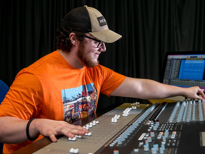 A young man in an orange t-shirt and baseball cap smiles as he makes adjustments to the dials and sliders on a mixer board.