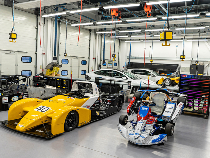A large well-lit garage with three racing cars in the foreground at different stages of assembly.