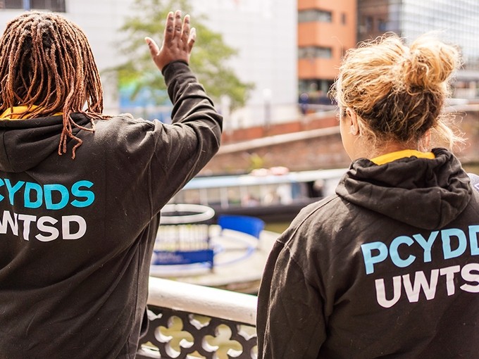 Mae dwy fenyw ifanc sy’n gwisgo hwdis PCYDDS yn chwifio tuag at gwch sy’n mynd drwy Gas Street Basin, Birmingham.