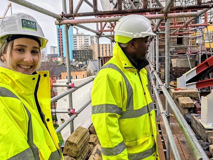 A man and a woman in hard hats and hi-vis jackets stand on a scaffolding walkway; the woman smiles to the camera while the man looks towards the building site.