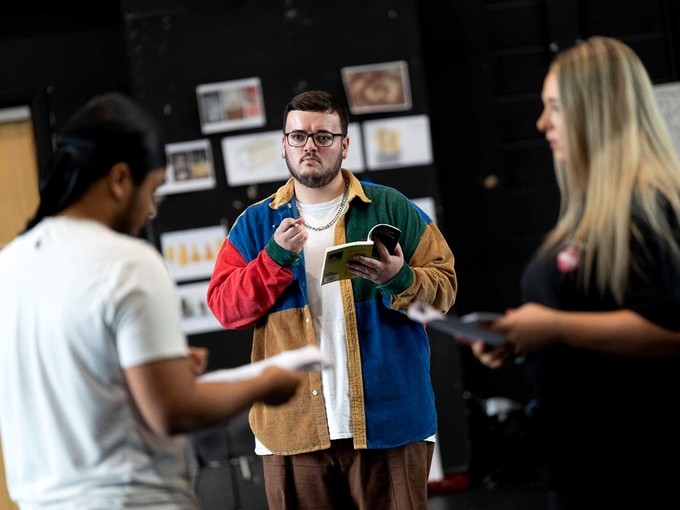 William Kingshott yn sefyll mewn ystafell ymarfer; mae’n dal sgript drama mewn un llaw ac mae ganddo bensil yn y llall; mae ei lygaid yn canolbwyntio ar fyfyrwraig arall.