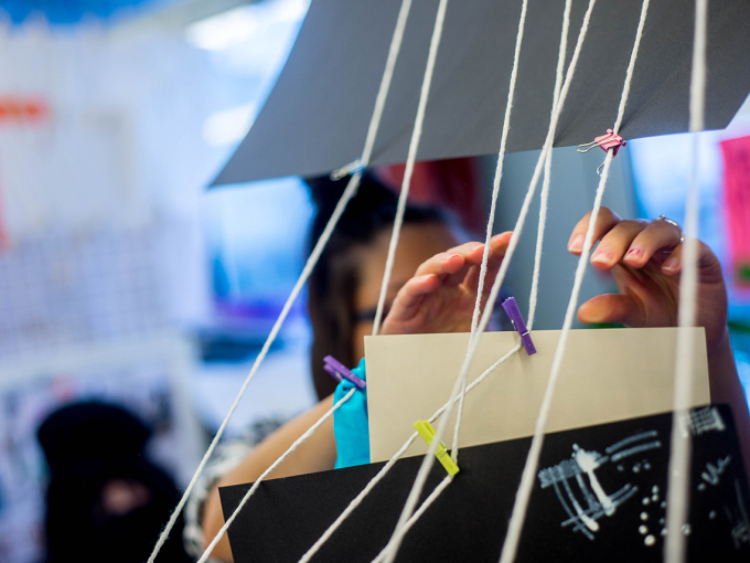 In the foreground, paper designs hang from strings in a classroom; an out-of-focus figure lifts her hands towards one of the suspended papers.