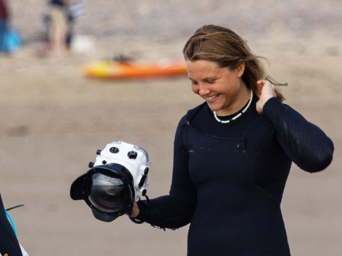 A person in a wetsuit smiling and walking on a beach while holding a camera