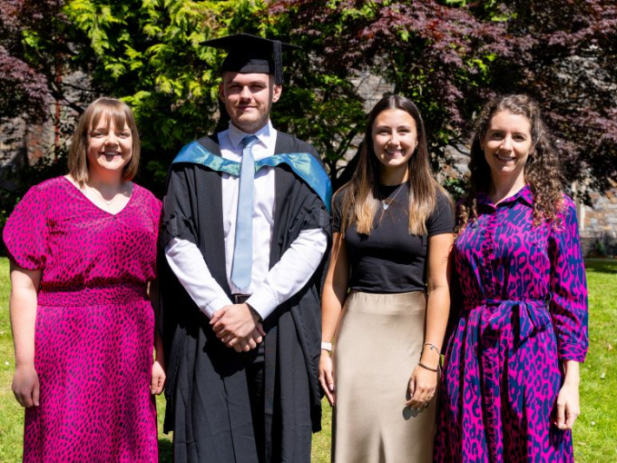 A group of four people standing on greenery. One of which is dressed in his cap and gown.